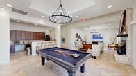 Located at 1101 Longhorn Rd in Fort Worth, TX, this single-floor clubhouse offers a well-appointed communal space designed for comfort, socializing, and productivity. The open-concept layout flows seamlessly across several distinct functional zones, all united by elegant herringbone-patterned tile flooring, crown molding, and recessed lighting throughout.

The living area serves as a central gathering point, anchored by a striking stone fireplace adorned with Western-inspired artwork. Comfortable patterned armchairs and a leather sofa surround the seating area, while large windows frame views of the outdoor pool and landscaping, flooding the space with natural light.

Adjacent to the lounge is a full-size billiards table positioned beneath a dramatic candelabra-style chandelier, with a wall-mounted flat-screen TV nearby for entertainment. The kitchen features rich dark wood cabinetry, granite countertops, stainless steel appliances including a full refrigerator and microwave, and a breakfast bar with seating — ideal for community gatherings. A dedicated Starbucks coffee station with granite counters adds a premium touch near the entrance.

A business center nook provides two iMac workstations set against artfully decorated walls, offering a quiet area for focused work. The overall ambiance is warm, contemporary, and thoughtfully curated with organic décor elements throughout.