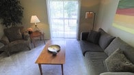 This single-floor residence offers a thoughtfully arranged living space designed for comfort and functionality. The living room serves as the heart of the home, featuring soft wall-to-wall carpeting that adds warmth underfoot. A large sliding glass door dominates one wall, flooding the room with natural light and providing a seamless connection to the outdoor area beyond, where lush greenery is visible. The space is well-appointed with a classic wood coffee table as a central focal point, complemented by a plush gray sofa and coordinating accent pillows. A wooden side table with a warm table lamp adds a cozy corner detail, while a floor lamp on the opposite side provides additional ambient lighting. The neutral wall tones and carpet create a calm, versatile backdrop that lends itself well to a variety of décor styles. The layout is practical and open, allowing for easy movement throughout the room. Overall, the space strikes a balance between simplicity and comfort, making it an inviting environment well-suited for both relaxing and everyday living.