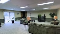 This single-floor space centers around a generously sized living area designed for comfort and functionality. The room features a two-tone wall treatment, with a deep sage green on the lower half and a lighter shade above, creating a grounded and cohesive aesthetic throughout. Recessed fluorescent panel lighting is evenly distributed across the ceiling, ensuring consistent, bright illumination throughout the space.

A standout architectural feature is the corner wood-burning stove, framed by a rustic dark wood surround that adds warmth and character to the room. The living area is thoughtfully arranged with plush, sage-toned sofas surrounding a dark coffee table, creating an inviting seating arrangement suited for both relaxation and social gatherings. A wall-mounted air conditioning unit provides climate control, while potted plants placed throughout the room introduce natural elements and a touch of greenery.

The space is finished with neutral carpeting that complements the earthy color palette, adding softness underfoot. An entertainment center with a media console anchors one wall, while a decorative clock and lamp contribute to the room's lived-in, welcoming ambiance. Overall, this space offers a practical and comfortable environment with a warm, casual character.