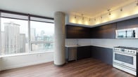 This contemporary high-rise unit delivers a sleek, urban living experience defined by clean lines and modern finishes throughout. The open-concept living and kitchen area is anchored by a striking exposed concrete structural column, which adds an industrial character to the space. The kitchen is outfitted with dark flat-panel cabinetry extending to the ceiling, bright white countertops, and a full suite of stainless steel appliances including a gas range, built-in microwave, French door refrigerator, and dishwasher. Track lighting mounted on a recessed upper cabinet shelf provides warm, directed illumination across the workspace. Rich hardwood flooring flows throughout the main living area, complementing the modern aesthetic. Floor-to-ceiling windows flood the space with natural light and frame sweeping city skyline views, bringing the urban landscape directly into the home. A stacked washer and dryer unit is conveniently integrated near the kitchen. The bedroom offers plush carpeting underfoot, crisp white walls, and a large window showcasing additional city views with a roller shade for privacy. The bathroom features a deep soaking tub surrounded by large-format, neutral-toned tile work, lending a clean and spa-like quality to the room.