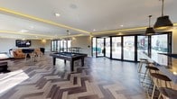 This single-floor amenity space is designed as a multifunctional social hub, combining a bar, lounge, and game room into one cohesive, open-concept layout. The interior is anchored by a striking full-service bar outfitted with a dramatic dark granite countertop featuring vivid blue and gold veining, complemented by warm wood paneling, leather bar stools, draft taps, and multiple wall-mounted televisions. Ambient LED cove lighting runs along the ceiling perimeter throughout the space, casting a warm golden glow that enhances the modern aesthetic.

The lounge area features generously sized sectional seating in tan leather and navy blue, arranged around a large wall-mounted TV. Floor-to-ceiling windows flood the space with natural light and provide open views of the surrounding greenery. The herringbone-patterned hardwood flooring adds a refined, geometric detail that flows throughout the space.

The game area includes a full-sized pool table and additional high-top seating near more mounted screens. Expansive folding glass doors at the far end of the room open directly to an outdoor patio with visible lounge furniture and shade structures, seamlessly extending the social space outdoors.