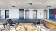 Located on the 9th floor of a modern residential building at 205 Rue De l'Équinoxe in Brossard, Quebec, this communal lounge offers a well-appointed and generously sized common living area designed for comfort and social interaction. The space is defined by an expansive curtain wall of floor-to-ceiling glass that wraps along one side, flooding the interior with abundant natural light and framing sweeping panoramic views of the surrounding cityscape and beyond. A glass door provides direct access to an outdoor terrace, seamlessly extending the lounge to the open air. The interior features a clean, contemporary design with a smooth white ceiling equipped with recessed lighting, complemented by durable carpeted flooring in a cool grey tone. The living area is furnished with a mix of leather sofas, mid-century modern armchairs in black, and accent chairs in a muted purple, all arranged around a solid wood coffee table atop a gold-and-ivory patterned area rug. A bank of three elevator doors is neatly integrated into the darker-toned accent wall, which is marked with a bold numeral "9," anchoring the space's identity as a floor-level amenity. Lush potted plants add a touch of greenery, enhancing the bright and welcoming ambiance of this thoughtfully designed communal retreat.