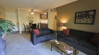This single-floor residence offers a thoughtfully arranged open-concept layout that seamlessly integrates the living, dining, and kitchen areas into one cohesive space. The living room serves as the central hub, featuring plush, dark upholstered seating accented with deep red throw pillows, complemented by a glass-top coffee table that adds a modern touch. Soft, neutral-toned carpeting runs throughout the space, contributing to a warm and comfortable ambiance. A ceiling fan with integrated lighting provides both airflow and illumination overhead, while a textured accent wall adds depth and visual interest to the room. Adjacent to the living area, a defined dining space accommodates a table and chairs, ideal for everyday meals or casual gatherings. The kitchen, visible in the background, is outfitted with dark cabinetry and a light countertop, offering a practical workspace. Warm ambient lighting enhances the overall inviting atmosphere throughout. The open sightlines between rooms create a sense of spaciousness and connectivity, making this a comfortable and functional single-level living space well-suited for everyday life.