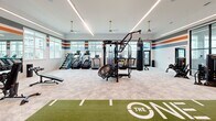 Located at 4415 Percival Rd in Columbia, SC, this expansive fitness center at The One at Columbia is thoughtfully designed to support a full range of training needs across two dedicated exercise rooms. The main workout area is a generously proportioned space featuring a signature branded turf training lane, a comprehensive selection of free weights and dumbbell racks, cable machines, multi-functional strength stations, and a heavy punching bag — accommodating everything from weightlifting to functional and combat training. Large windows line the walls, flooding the space with natural light and providing views of the surrounding community. Bold color-block striping in orange, red, and blue accents the upper walls, lending the space a dynamic, energetic aesthetic. Sleek linear LED lighting and ceiling fans keep the environment bright and comfortable. A wall of floor-to-ceiling black-framed glass panels visually connects the two rooms, creating an open, airy feel. The second exercise room features light hardwood flooring, additional strength and resistance equipment, stability balls, TRX suspension trainers, and dedicated cardio machines including treadmills and stair climbers, all bathed in natural light from oversized windows. The overall design is modern and motivating, purpose-built to deliver a premium fitness experience.