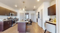 This single-floor unit features a contemporary open-concept design centered around a well-appointed kitchen. The kitchen is equipped with dark espresso cabinetry, granite countertops, and a full suite of stainless steel appliances, including a refrigerator, range, and microwave. A substantial kitchen island with matching granite surface provides additional prep space and doubles as a casual dining area, accommodating multiple bar-height stools beneath pendant lighting. Recessed lighting throughout the kitchen and adjoining living area ensures consistent illumination across the space.

Light-toned hardwood-style flooring runs through the main living and kitchen areas, contributing to a clean, cohesive aesthetic. The white walls and open layout create a bright, airy atmosphere. The bedroom offers plush carpeting, natural light through a large window dressed with dark curtains and horizontal blinds, and a calm, neutral color palette.

The bathroom features dark wood cabinetry with brushed nickel hardware, a granite vanity with an undermount sink, a large mirror, and a full shower-tub combo. Hardwood-style flooring continues into the bathroom, maintaining visual continuity throughout the unit. The overall design balances modern finishes with functional, everyday livability.