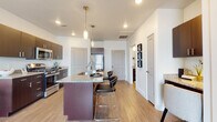 This single-floor unit features a contemporary open-concept design centered around a well-appointed kitchen. The kitchen is equipped with dark espresso cabinetry, granite countertops, and a full suite of stainless steel appliances, including a refrigerator, range, and microwave. A substantial kitchen island with matching granite surface provides additional prep space and doubles as a casual dining area, accommodating multiple bar-height stools beneath pendant lighting. Recessed lighting throughout the kitchen and adjoining living area ensures consistent illumination across the space.

Light-toned hardwood-style flooring runs through the main living and kitchen areas, contributing to a clean, cohesive aesthetic. The white walls and open layout create a bright, airy atmosphere. The bedroom offers plush carpeting, natural light through a large window dressed with dark curtains and horizontal blinds, and a calm, neutral color palette.

The bathroom features dark wood cabinetry with brushed nickel hardware, a granite vanity with an undermount sink, a large mirror, and a full shower-tub combo. Hardwood-style flooring continues into the bathroom, maintaining visual continuity throughout the unit. The overall design balances modern finishes with functional, everyday livability.