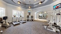 Located at 909 Penn Waller Rd in Savannah, Georgia, this fitness center offers a well-designed workout environment that combines functionality with refined architectural details. The space is organized to accommodate both strength training and cardiovascular exercise, with a comprehensive selection of cable machines, weight benches, treadmills, ellipticals, and stationary bikes distributed throughout the room.

Architecturally, the facility stands out with its striking multi-tiered tray ceilings trimmed in gray and white crown molding, lending a sense of elevated height and sophistication uncommon in fitness spaces. Pendant light fixtures and recessed lighting provide consistent, well-distributed illumination throughout. A signature arched entryway connects the two main zones of the fitness center, framed by full-length mirrors that visually expand the space and assist with workout form.

The floors are covered in durable, patterned carpet designed for gym use, while large windows along the perimeter invite abundant natural light and offer views of the surrounding greenery. Wall-mounted flat-screen TVs are positioned for easy viewing from multiple equipment stations. The soft gray-and-white color palette, clean white baseboards, and polished finishes create an environment that feels both professional and welcoming, making this fitness center a standout amenity within its community.