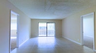 This single-floor residence offers a practical and well-organized layout designed to maximize living comfort. The floor plan reveals a central living area flanked by two spacious rooms on either side, creating a balanced and symmetrical arrangement. A dedicated area toward the rear of the unit accommodates a kitchen and additional functional spaces, while compact rooms near the front appear to serve as bathrooms or utility areas.

The living area is a standout feature of this home, finished with large, light-colored ceramic tile flooring that reflects natural light and adds a clean, airy feel to the space. A textured ceiling adds a classic touch overhead, while warm, neutral-toned walls contribute to a welcoming atmosphere. A sliding glass door provides direct access to an outdoor balcony or patio, allowing natural light to flood the interior and offering a seamless connection to the outdoors.

The two flanking rooms are generously proportioned and share a similar aesthetic, with tile flooring and neutral walls ready to accommodate a variety of uses. Overall, the space presents a straightforward and functional design, with an open central area ideal for everyday living and entertaining.