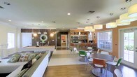 This single-floor community amenity space is thoughtfully designed to accommodate a variety of social and recreational activities. The open-concept layout seamlessly connects several distinct zones, creating a cohesive and inviting atmosphere throughout.

Upon entering, a lounge area greets visitors with comfortable seating, including a plush sofa adorned with decorative pillows, set against a richly textured, dark feature wall with stylish wall décor. Adjacent to the lounge sits a pool table, making this section an ideal spot for casual recreation and entertainment.

The dining area occupies the central and right portions of the space, featuring multiple tables and a mix of colorful modern chairs in shades of green, orange, and white — adding a lively, contemporary feel. Large windows with white blinds allow natural light to fill this section of the room.

Toward the back, a well-appointed kitchenette serves the space with counter seating at a bar-height island, open shelving stocked with supplies, a microwave, a toaster oven, and a coffee maker — offering convenient self-service amenities for residents or guests.

Recessed lighting and pendant fixtures illuminate the entire space warmly, while wood-look and tile flooring ground the design with a clean, durable finish. The overall ambiance is energetic yet comfortable, suited for community gatherings and everyday use.