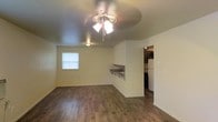 This single-floor space features an open layout that seamlessly connects the main living area to the kitchen. The living area is finished with warm hardwood-style flooring that extends throughout, contributing to a cohesive and clean aesthetic. A ceiling fan with an integrated light fixture serves as the central lighting source, providing both functionality and a modest focal point overhead.

A window along one wall allows natural light to enter the living space, brightening the room and creating an airy feel. The walls are finished in a neutral tone, offering a versatile backdrop suitable for a variety of interior styles.

A built-in bar-style counter with support brackets is positioned along the wall between the living area and kitchen, providing a convenient pass-through or casual dining surface. This feature enhances the connectivity between the two spaces.

The kitchen, visible through an open doorway, includes dark cabinetry and a white refrigerator, with overhead lighting illuminating the area. The layout appears efficient and functional, making practical use of the available space. Overall, the unit presents a straightforward, functional design with warm finishes throughout.