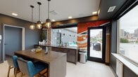 This contemporary single-floor amenity space combines bold design with practical functionality, creating an inviting environment for gathering and socializing. The interior is anchored by a striking kitchen area outfitted with dark cabinetry, a stone-topped island with seating for multiple guests, and a cluster of designer pendant lights overhead. A large pass-through window connects the kitchen to an adjoining covered outdoor terrace, which features outdoor seating and a grill, seamlessly blending indoor and outdoor living.

A standout visual element throughout the space is a dramatic swirling orange, red, and white mural column, adding artistic energy to the room. Complementing this, textured stone-patterned wall panels and curated decorative accents contribute to the overall upscale aesthetic. The flooring consists of large-format light tile throughout, providing a clean, cohesive look.

A dedicated lounge and dining area features a built-in banquette upholstered in soft gray, paired with round marble-topped tables and mixed leather chairs beneath a cascading array of globe pendant lights set against a deep navy accent wall. Expansive windows frame broad city and skyline views, filling the space with natural light. The overall ambiance is modern, sophisticated, and thoughtfully designed for communal use.