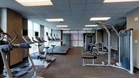Located at 3737 Chestnut Street in Philadelphia, PA, this contemporary fitness center offers a well-equipped, single-floor workout facility designed to support a full range of exercise routines. The space is thoughtfully laid out to maximize usability and flow, with dedicated zones for both cardiovascular and strength training activities. A row of treadmills and stationary bikes lines the perimeter near expansive floor-to-ceiling windows, flooding the space with natural light and providing inspiring views of the surrounding cityscape. The strength training area features a multi-station cable and weight machine setup, complemented by additional free weight and resistance equipment. Patterned low-pile carpet tiles cover the flooring throughout, adding visual interest while providing a functional surface underfoot. A dropped grid ceiling with recessed panel lighting ensures consistent, even illumination across the entire space. Glass partitions and dark accent walls with textured brick detailing create visual separation and add an urban, modern aesthetic to the interior. The overall ambiance is clean, professional, and energizing — designed to motivate and accommodate residents or guests seeking a convenient, fully equipped fitness experience.