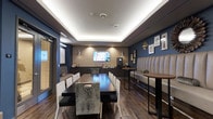 This coffee lounge is a thoughtfully designed social space that combines comfort and contemporary style. The room is anchored by a tray ceiling with integrated LED cove lighting, which casts a warm, ambient glow throughout the space and lends a polished, upscale feel. Rich hardwood-style flooring runs the length of the lounge, complementing the deep navy blue and warm taupe accent walls that give the room a sophisticated, welcoming atmosphere.

Seating options are varied and plentiful. A long, dark communal dining table with an eclectic mix of white, black, and upholstered chairs occupies the central area, making it well-suited for group gatherings or collaborative work. Along one wall, a generously sized, channel-tufted cream banquette provides comfortable lounge-style seating paired with round wood-top bistro tables — perfect for more intimate conversations or quiet relaxation.

A flat-screen television is mounted prominently on the far accent wall, adding an entertainment element to the space. Adjacent to it, a dedicated beverage station is equipped with a coffee machine and counter space for refreshments. The walls are further accented with a curated gallery of framed artwork and decorative mirrors, adding character and visual depth. Pendant lighting fixtures contribute additional warmth, completing this inviting and well-appointed coffee lounge.