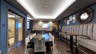 This coffee lounge is a thoughtfully designed social space that combines comfort and contemporary style. The room is anchored by a tray ceiling with integrated LED cove lighting, which casts a warm, ambient glow throughout the space and lends a polished, upscale feel. Rich hardwood-style flooring runs the length of the lounge, complementing the deep navy blue and warm taupe accent walls that give the room a sophisticated, welcoming atmosphere.

Seating options are varied and plentiful. A long, dark communal dining table with an eclectic mix of white, black, and upholstered chairs occupies the central area, making it well-suited for group gatherings or collaborative work. Along one wall, a generously sized, channel-tufted cream banquette provides comfortable lounge-style seating paired with round wood-top bistro tables — perfect for more intimate conversations or quiet relaxation.

A flat-screen television is mounted prominently on the far accent wall, adding an entertainment element to the space. Adjacent to it, a dedicated beverage station is equipped with a coffee machine and counter space for refreshments. The walls are further accented with a curated gallery of framed artwork and decorative mirrors, adding character and visual depth. Pendant lighting fixtures contribute additional warmth, completing this inviting and well-appointed coffee lounge.