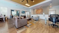 Located at 1100 Townsend Blvd in Fredericksburg, Virginia, this single-floor community clubhouse offers a well-appointed and thoughtfully designed gathering space. The main lounge area immediately draws attention with its soaring tray ceiling accented by a gold finish, a large ceiling fan, and a skylight that floods the room with natural light. Wood-look flooring runs throughout, lending warmth and continuity to the space.

The lounge is furnished with plush, neutral-toned sofas and accent chairs arranged around a bold geometric area rug featuring shades of orange, gray, and tan — a color palette echoed in the striking orange square lattice wall sculpture that serves as a focal point on the main wall. A wall-mounted flat-screen television and a dark wood media console complete the entertainment area, while multiple French-style windows and glass doors invite views of the surrounding greenery.

A dedicated seating nook features high-top bar-height seating at a round table positioned in front of large windows, offering an abundance of natural light and outdoor views. Nearby, a coffee bar area with a shiplap-accented wall, built-in shelving, and navy blue upholstered bar stools provides a casual spot for refreshments. The overall ambiance is modern and inviting, blending contemporary furnishings with warm architectural details.