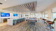 Located at 1250 Marden Rd in Apopka, Florida, this thoughtfully designed community amenity space offers a vibrant coastal aesthetic across a generously sized single-floor layout. The open-plan interior is anchored by a striking long bar island clad in distressed teal-toned paneling and topped with a crisp white quartz surface, lined with warm wood-and-metal bar stools. Pendant lights hang above the bar in a row, complementing the eye-catching slatted wood ceiling treatment that adds texture and a resort-like warmth overhead. Dual flat-screen televisions are mounted behind the bar area, which also features a wet bar and wine refrigerator, making it ideal for social gatherings. A full-sized billiards table occupies the central lounge zone, surrounded by patterned carpet with green and blue accents. Large windows along the perimeter flood the space with natural light and offer outdoor views, while a balcony-accessible area provides additional seating for smaller groups. A separate conference-style room accommodates private meetings or events, furnished with a long communal table, mid-century modern leather chairs, and elegant gold cube pendant lighting, adorned with framed palm tree artwork. Light hardwood flooring connects the transitional hallway areas, tying the entire space together with a clean, cohesive look.