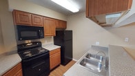 This single-floor space offers a practical and well-organized layout designed for comfortable everyday living. The kitchen serves as a central highlight, showcasing warm honey-toned wood cabinetry that provides generous storage with both upper and lower cabinet configurations. Black appliances — including an electric range, over-the-range microwave, and a full-size refrigerator — create a cohesive, modern aesthetic against the neutral beige walls. The countertops feature a speckled laminate surface that balances durability with style, complemented by a double-basin stainless steel sink fitted with a single-handle faucet. The kitchen flooring consists of light wood-tone planks that add warmth and are easy to maintain. A flush-mounted overhead light fixture provides bright, even illumination throughout the cooking area. The textured ceiling adds a classic touch consistent throughout the space. The overall ambiance is functional and inviting, with a neutral color palette that offers a versatile backdrop for personalization. This space is thoughtfully designed to maximize utility while maintaining a clean, cohesive look throughout.
