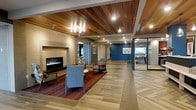 This single-floor space presents a thoughtfully designed lobby that balances warmth and contemporary style. The ceiling is clad in rich, horizontal wood planks, lending a natural warmth that contrasts beautifully with the smooth, large-format tile flooring arranged in a herringbone pattern. Recessed lighting and a chandelier-style fixture illuminate the space evenly, highlighting the refined finishes throughout.

A central seating area anchors the lobby, featuring mid-century modern armchairs in muted blue tones arranged around a low table atop a deep burgundy area rug. A sleek, wall-mounted linear fireplace serves as a focal point, framed in neutral stone-toned tile that extends to the surrounding wall, creating an inviting atmosphere.

The space opens into wider corridors that lead to additional rooms, including what appears to be a reception or service area enclosed in glass, offering both transparency and separation. Accent walls painted in deep teal blue add depth and visual interest, while framed artwork contributes to a curated, professional ambiance.

The open layout and generous sightlines create a sense of spaciousness and ease of navigation. Overall, the design strikes a balance between functional, welcoming, and aesthetically polished — well-suited for a professional or hospitality-oriented environment.