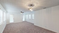 This single-story space is designed with a practical, open layout that connects the main living and dining areas seamlessly. The living room is generously sized, finished with neutral-toned carpet and crisp white walls that create a bright, clean atmosphere. A decorative pendant light cluster serves as a central focal point overhead, while a ceiling fan provides additional airflow. Multiple windows fitted with white blinds line the exterior wall, allowing natural light to filter through and illuminate the space. A door provides direct exterior access from this area.

The kitchen is outfitted with raised-panel gray cabinetry offering substantial storage above and below the counters, complemented by white laminate countertops and white appliances including a coil-burner range and a full-size refrigerator. Dark wood-look flooring grounds the kitchen, providing a clear visual distinction from the carpeted areas.

The bedrooms continue the neutral carpet and white wall palette, each equipped with ceiling fans and windows dressed with blinds for natural light and privacy. The bathroom features a wide vanity with a single undermount sink, light countertops, white cabinetry, and a large wall mirror. Throughout, textured popcorn ceilings are consistent across all rooms.