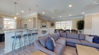 This single-story farmhouse features an open, flowing layout that seamlessly connects the kitchen, living, and dining areas. Light hardwood-style flooring runs throughout, complementing the warm, neutral wall tones and crisp white crown molding and beadboard wainscoting that add classic architectural character to each space.

The kitchen is well-appointed with light wood cabinetry, light stone countertops, black appliances including a range, refrigerator, and dishwasher, and a window above the sink that draws in natural light. Pendant lighting accents the space, and a breakfast bar with seating provides a casual dining option.

The open living area features a large sectional sofa, a wall-mounted television, and a ceiling fan, creating a comfortable and functional gathering space. Beadboard wainscoting lines the lower walls, and large multi-pane windows flood the room with natural light while offering views of the pool and surrounding outdoor area.

The dining area accommodates a generously sized dark wood table with ample seating, positioned beside expansive windows that frame views of the landscaped exterior. Throughout the farmhouse, the combination of natural light, classic detailing, and cohesive design creates a welcoming and well-organized communal environment.