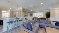 This single-story farmhouse features an open, flowing layout that seamlessly connects the kitchen, living, and dining areas. Light hardwood-style flooring runs throughout, complementing the warm, neutral wall tones and crisp white crown molding and beadboard wainscoting that add classic architectural character to each space.

The kitchen is well-appointed with light wood cabinetry, light stone countertops, black appliances including a range, refrigerator, and dishwasher, and a window above the sink that draws in natural light. Pendant lighting accents the space, and a breakfast bar with seating provides a casual dining option.

The open living area features a large sectional sofa, a wall-mounted television, and a ceiling fan, creating a comfortable and functional gathering space. Beadboard wainscoting lines the lower walls, and large multi-pane windows flood the room with natural light while offering views of the pool and surrounding outdoor area.

The dining area accommodates a generously sized dark wood table with ample seating, positioned beside expansive windows that frame views of the landscaped exterior. Throughout the farmhouse, the combination of natural light, classic detailing, and cohesive design creates a welcoming and well-organized communal environment.
