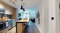 This thoughtfully designed single-floor residence showcases a clean, contemporary aesthetic throughout its open-concept layout. The kitchen is outfitted with light wood cabinetry, stainless steel appliances including a range, microwave, and dishwasher, paired with a textured tile backsplash and light stone countertops. A sleek kitchen island with bar seating and striking dual pendant lights serves as the centerpiece connecting the kitchen to the dining and living areas. Wide-plank hardwood flooring flows throughout, lending warmth and continuity across the space. The living area is anchored by a large sectional sofa and features a standout city map wall mural, adding a bold artistic focal point to the room. Large floor-to-ceiling windows flood the living and bedroom areas with natural light while framing urban skyline views. Two well-proportioned bedrooms each offer generous window space, warm wood flooring, and easy access to natural light. The bathroom is finished with light wood vanity cabinetry, a stone-toned countertop, full tub-shower combination with white subway tile surround, and tasteful framed artwork. Throughout, the residence maintains a cohesive modern design palette of neutral tones, natural wood textures, and carefully curated details.