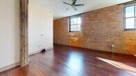 Located at 427 Pershing Ave in Davenport, Iowa, this one-bedroom, two-bathroom unit blends industrial character with modern comfort. Rich hardwood flooring flows throughout the main living areas, complemented by striking exposed brick accent walls that lend an authentic, loft-inspired aesthetic. The open-concept living and kitchen area is anchored by a well-equipped kitchen featuring warm wood cabinetry, granite countertops, stainless steel appliances including a range, microwave, dishwasher, and refrigerator, and tile flooring. Track lighting and an exposed wooden beam overhead add to the space's industrial appeal. Large black-framed windows invite natural light and frame views of the surrounding area. The living room offers generous proportions with a ceiling fan and additional exposed brick, creating a warm and inviting atmosphere. The primary bedroom is a comfortable retreat, also featuring hardwood floors and a signature exposed brick wall. The two bathrooms are tastefully finished with granite-topped vanities, tile flooring, and clean white surrounds. One bathroom conveniently includes an in-unit washer and dryer. A hallway with closet completes the thoughtful layout of this character-rich Davenport residence.