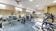 Located in Santa Cruz, California, this fitness center offers a thoughtfully designed workout space that accommodates a comprehensive range of exercise needs. The room features a drop-panel ceiling with recessed lighting panels and ceiling fans that ensure consistent airflow and a bright, comfortable environment throughout. A row of clerestory windows along the upper walls allows natural light to filter in, lending an open and airy feel to the space. Full-length mirrors along one wall visually expand the room while providing functional feedback during workouts. The flooring is a standout design element, featuring a geometric chevron pattern in shades of blue, gold, and neutral tones that adds a modern, dynamic aesthetic underfoot. The equipment layout is well-organized, with cardio machines — including treadmills, stationary bikes, and ellipticals — positioned alongside a variety of strength training machines and cable systems. A wall-mounted flat-screen TV is available for entertainment during workouts. An adjacent alcove provides a water fountain station for easy hydration. Decorative artwork adorns the walls, adding a touch of personality to the functional space. The overall ambiance is clean, energetic, and welcoming, making it a capable and well-appointed fitness facility.