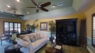 This single-story clubhouse offers a comfortable and inviting communal lounge space designed for relaxation and socializing. The main gathering area is anchored by a plush, cream-colored sofa dressed with decorative striped throw pillows, positioned to face a substantial dark wood entertainment center housing a large flat-screen television. The rich, espresso-toned cabinetry features ornate detailing and ample storage, serving as a striking focal point in the room.

Warm yellow walls, accented by a contrasting sage green tray ceiling, create a cheerful and welcoming atmosphere. Recessed lighting and a ceiling fan with a brushed nickel finish provide both illumination and comfort throughout the space. Lush tropical potted plants are thoughtfully placed throughout the room, adding a vibrant, natural element to the interior.

A decorative framed artwork piece adorns the wall, complementing the room's warm color palette. An ornate, patterned area rug anchors the seating arrangement, while a dark wood coffee table with a glass top completes the gathering space. Overall, the clubhouse lounge exudes a relaxed yet polished ambiance ideal for community gatherings.