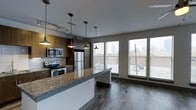 This single-floor space showcases a thoughtfully designed open-concept layout that seamlessly connects the kitchen and living areas. The kitchen is a standout feature, equipped with stainless steel appliances including a refrigerator, electric range, and built-in microwave. Rich, dark wood cabinetry provides ample storage while contributing to the space's contemporary aesthetic. Granite countertops with warm tonal variations extend across the kitchen surfaces, including a generous island or peninsula that serves as both a functional workspace and a natural gathering point.

A series of industrial-style pendant lights hang at varying heights above the counter, adding character and focused illumination. Track lighting along the kitchen ceiling provides additional brightness. The flooring throughout is a durable wood-look material in a cool gray tone, tying the space together cohesively.

Large floor-to-ceiling windows along one wall flood the living area with natural light and frame views of a city skyline, suggesting an elevated urban location. Horizontal blinds offer light control and privacy. The overall ambiance is modern and urban, balancing functional design with contemporary finishes.