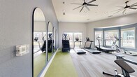 Located at 529 Barker-Clodine Road in Houston, TX, this fitness center offers a thoughtfully designed exercise space built for effective and comfortable workouts. The room is finished in a cool, calming palette of soft blues and neutral grays, creating an energizing yet focused atmosphere. Patterned, striped carpet tiles cover the main floor area, while a stretch of green flooring along the accent wall defines a dedicated stretching or functional training zone. A standout design feature is the series of elegant arched mirrors mounted along the blue accent wall, which visually expand the space and allow users to monitor their form. Large windows and a bay window area flood the room with natural light and offer pleasant outdoor views, enhancing the overall workout experience. Overhead, large ceiling fans with recessed lighting ensure consistent air circulation throughout. The facility is equipped with a comprehensive selection of cardio machines, including True-brand treadmills, ellipticals, upright and recumbent bikes, stair climbers, and rowing machines, providing a full range of cardiovascular training options. The space is accessible via a dedicated entry door, ensuring a private and convenient fitness environment.