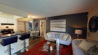 This single-floor residence offers a thoughtfully designed layout that balances comfort and contemporary style throughout. The living room serves as the heart of the home, featuring wood-style flooring, a bold dark accent wall adorned with large-format artwork, and tufted sofas centered on a vibrant red area rug. A glass sliding door floods the space with natural light and opens directly onto a private patio, providing seamless indoor-outdoor connectivity.

Adjacent to the living area, the dining space is served by a pendant light fixture, while the galley-style kitchen features white cabinetry with arched detailing, dark granite-style countertops, and black appliances, including a full-size refrigerator and range.

The residence includes two bedrooms, each with plush carpeting and ceiling fans. The primary bedroom features a dramatic navy accent wall with botanical artwork, while the second bedroom offers a bright window with outdoor views and convenient closet access.

Two full bathrooms complement the bedrooms. Both feature white cabinetry, dark speckled countertops, large vanity mirrors with overhead lighting, and tub-shower combinations with tile surrounds and wood-style flooring, providing a clean, functional finish throughout.