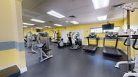 This single-level fitness center is designed to accommodate a comprehensive range of workout routines, offering both cardio and strength training areas within a spacious, open layout. The interior is immediately distinguished by its bold yellow walls accented with a gray horizontal stripe, creating an energetic and motivating atmosphere. Durable dark rubber flooring covers the entire space, providing cushioning and traction suitable for high-intensity exercise.

The cardio zone is well-stocked with multiple treadmills, elliptical trainers, and a rowing machine, several of which are equipped with individual digital consoles. Ceiling fans and portable floor fans supplement the ventilation system, ensuring comfort during use. Wall-mounted televisions are positioned for easy viewing from the cardio equipment.

The strength training area features a multi-station cable machine, a squat/weight rack, adjustable benches, and a full dumbbell rack with weights organized along a mirrored wall. Suspension training straps and additional resistance accessories are mounted to the walls for functional fitness exercises. Mirrors along the strength section enhance the sense of space and allow users to monitor their form.

Natural light enters through windows on multiple walls, while a drop ceiling fitted with recessed fluorescent panels ensures consistent, even illumination throughout the facility.