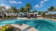 Located in Royal Palm Beach, Florida, this outdoor amenity space offers a quintessential South Florida resort experience centered around a generously sized freeform swimming pool with crystal-clear, blue-tinted water. The pool's curved, organic shape invites leisurely swims and relaxation, while its surrounding paver deck provides ample space for sunbathing and socializing. A charming poolside cabana structure serves as the visual focal point, featuring an open-air design with a covered roof, an outdoor television, and vibrant blue lounge chairs arranged for comfort and conversation. Colorful tropical plantings, including vivid red and pink flowers, line the cabana and deck areas, adding bursts of natural color throughout. Towering royal palms and mature shade trees frame the entire space, creating a lush, tropical canopy that evokes a sense of a private oasis. Elegant white shade canopies and additional seating areas are thoughtfully positioned around the perimeter, offering shaded retreats from the Florida sun. The overall ambiance is bright, inviting, and distinctly tropical, making this outdoor space an ideal setting for relaxation and recreation in a beautifully maintained community environment.