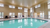 This property offers two distinct pool environments designed to accommodate year-round aquatic recreation. The indoor pool is housed within a generously sized, single-story structure featuring soaring ceilings and an abundance of natural light streaming through large, multi-pane windows positioned at multiple heights along the walls. The warm, neutral-toned interior is finished with tiled accents along the pool surround, and the space includes accessible restroom facilities, lounge chairs, and poolside seating. The expansive clear-water pool is complemented by an adjacent spa, and the open layout provides ample deck space for relaxation.

The outdoor pool area presents a spacious concrete deck surrounding a large rectangular pool with clear blue water. A separate spa is positioned adjacent to the pool. Lounge chairs and umbrella-shaded seating are arranged along the deck, offering comfortable spaces to unwind. The pool is accessed through a dedicated pool house featuring rustic wood-framed doors, stucco exterior walls, and posted pool rules signage. Mature trees and lush landscaping frame the perimeter, providing shade and a pleasant outdoor ambiance set against the backdrop of a multi-story brick residential building.
