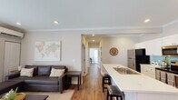 This thoughtfully designed 2-bedroom, 2-bathroom residence offers a comfortable and functional single-floor layout with over 1,150 square feet of living space. The open-concept living and kitchen area serves as the heart of the home, featuring warm hardwood-style flooring, crown molding, and recessed lighting throughout. The kitchen is equipped with stainless steel appliances, crisp white shaker cabinetry, a teal tile backsplash, and a quartz-topped island with bar seating — ideal for casual dining and entertaining. A dedicated dining area sits adjacent to the kitchen, while the living room provides a relaxed space accented by a large world map wall art piece and a dark leather sectional sofa. Each bedroom is generously sized, filled with natural light from large windows, and features its own wall-mounted mini-split HVAC unit for individual climate control. Both bathrooms are finished with white cabinetry, clean tile work, and hardwood-style flooring, with one featuring a full shower. A central hallway connects all rooms efficiently, contributing to a well-organized and cohesive floor plan. The overall aesthetic is modern yet warm, with neutral tones and tasteful décor creating an inviting atmosphere throughout.