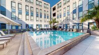 Located in New Orleans, LA, this outdoor pool and clubhouse amenity offers a well-appointed retreat designed for relaxation and social gatherings. The centerpiece is a generously sized rectangular swimming pool with crystal-clear blue water, elegantly framed by a wide tiled pool deck. The deck is lined with cushioned lounge chairs and shaded by large patio umbrellas, providing ample space for sunbathing and unwinding.

The pool area is nestled within a modern courtyard, enclosed on multiple sides by a contemporary multi-story building featuring a neutral facade with bold blue accent panels and expansive windows. Tall palm trees and lush tropical plantings, including oversized leafy plants in sleek rectangular planters, add a vibrant, resort-like atmosphere to the space. Outdoor seating areas with tables and chairs are positioned along the pool deck for casual dining or conversation.

Access to the Pool Clubhouse is provided via glass-paneled doors directly off the pool deck. The interior clubhouse features warm wood-toned finishes, tile flooring, pendant lighting, a white countertop bar with seating, and comfortable lounge furniture — creating a stylish indoor complement to the outdoor amenity. The entry vestibule serves as a transition space between the pool area and the clubhouse interior, complete with pool rules signage and a small utility station.