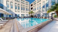 Located in New Orleans, LA, this outdoor pool and clubhouse amenity offers a well-appointed retreat designed for relaxation and social gatherings. The centerpiece is a generously sized rectangular swimming pool with crystal-clear blue water, elegantly framed by a wide tiled pool deck. The deck is lined with cushioned lounge chairs and shaded by large patio umbrellas, providing ample space for sunbathing and unwinding.

The pool area is nestled within a modern courtyard, enclosed on multiple sides by a contemporary multi-story building featuring a neutral facade with bold blue accent panels and expansive windows. Tall palm trees and lush tropical plantings, including oversized leafy plants in sleek rectangular planters, add a vibrant, resort-like atmosphere to the space. Outdoor seating areas with tables and chairs are positioned along the pool deck for casual dining or conversation.

Access to the Pool Clubhouse is provided via glass-paneled doors directly off the pool deck. The interior clubhouse features warm wood-toned finishes, tile flooring, pendant lighting, a white countertop bar with seating, and comfortable lounge furniture — creating a stylish indoor complement to the outdoor amenity. The entry vestibule serves as a transition space between the pool area and the clubhouse interior, complete with pool rules signage and a small utility station.