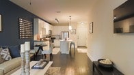 This single-floor residence offers a thoughtfully designed open-concept layout that seamlessly connects the living, dining, and kitchen areas into one cohesive and functional space. Rich, dark hardwood flooring runs throughout, grounding the interior with warmth and elegance.

The kitchen is a standout feature, outfitted with crisp white cabinetry, stainless steel appliances, and a central island with bar seating — ideal for casual dining or entertaining. A stylish pendant light hangs above the island, adding a refined focal point, while track lighting overhead provides practical illumination. The kitchen's clean white palette is beautifully contrasted by a bold dark accent wall that adds depth and visual interest to the open space.

The dining area, positioned adjacent to the kitchen, comfortably accommodates a small table and chairs, creating an intimate setting for meals. The living area flows naturally from the dining space, providing a relaxed and inviting environment.

A welcoming entryway with a classic white door and neutral walls completes the layout, offering a clean transition into the home. The overall ambiance is modern yet warm, with a careful balance of light tones and bold contrasts that give the space a stylish, curated character throughout.