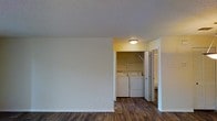 This single-floor space presents a straightforward and functional layout designed for everyday comfort. The main living area features rich, dark hardwood-style flooring that extends throughout, adding warmth and a sense of continuity to the space. The walls are finished in a neutral off-white tone, complemented by a textured popcorn ceiling that adds subtle dimension overhead.

A standout feature is the dedicated in-unit laundry area, tucked neatly behind bifold doors and equipped with a full-sized washer and dryer. Above the machines, built-in shelving provides convenient storage for laundry essentials. A ceiling-mounted light fixture illuminates the alcove, keeping the area bright and practical.

Adjacent to the laundry space, a doorway leads to an additional room, suggesting a multi-room configuration that supports versatile living arrangements. The open wall space in the main area provides ample room for flexible furniture placement. Overall, the space maintains a clean, understated aesthetic with functional details that prioritize ease of living.