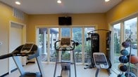 This fitness room is thoughtfully designed to support a comprehensive workout routine within a bright and welcoming environment. The space is outfitted with an array of professional-grade exercise equipment, including treadmills, an elliptical trainer, a stationary cycle, and a multi-function cable machine, providing options for both cardio and strength training. Large sliding glass doors span the exterior wall, flooding the room with natural light and offering unobstructed views of a beautifully landscaped outdoor patio. The patio beyond features stately white columns and lush tropical greenery, creating a serene and motivating backdrop during workouts. A wall-mounted flat-screen television is positioned prominently above the windows, allowing for entertainment while exercising. The walls are painted in a warm yellow tone, contributing to an energetic yet comfortable ambiance. Recessed ceiling lighting supplements the abundant natural light, ensuring the space remains well-lit at any time of day. The light-colored flooring is easy to maintain and complements the overall clean, airy aesthetic of the room. This fitness space effectively combines functionality with an inviting atmosphere, making it an ideal setting for daily exercise.