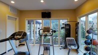 This fitness room is thoughtfully designed to support a comprehensive workout routine within a bright and welcoming environment. The space is outfitted with an array of professional-grade exercise equipment, including treadmills, an elliptical trainer, a stationary cycle, and a multi-function cable machine, providing options for both cardio and strength training. Large sliding glass doors span the exterior wall, flooding the room with natural light and offering unobstructed views of a beautifully landscaped outdoor patio. The patio beyond features stately white columns and lush tropical greenery, creating a serene and motivating backdrop during workouts. A wall-mounted flat-screen television is positioned prominently above the windows, allowing for entertainment while exercising. The walls are painted in a warm yellow tone, contributing to an energetic yet comfortable ambiance. Recessed ceiling lighting supplements the abundant natural light, ensuring the space remains well-lit at any time of day. The light-colored flooring is easy to maintain and complements the overall clean, airy aesthetic of the room. This fitness space effectively combines functionality with an inviting atmosphere, making it an ideal setting for daily exercise.