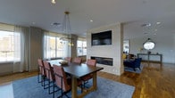 This single-floor space showcases a thoughtfully designed open-plan layout that seamlessly connects a dining area and a lounge, creating a versatile and inviting environment. The dining area is anchored by a substantial dark-wood table surrounded by upholstered rust-toned chairs, set atop a plush blue-gray area rug that adds texture and warmth to the hardwood flooring beneath. Above the table, a striking brass cluster pendant light fixture serves as a focal point, lending an elegant, contemporary touch to the space. Natural light flows in generously through large windows dressed with sheer curtains, offering views to the outside and brightening the neutral color palette of the walls and ceiling. A sleek, floor-level linear fireplace is built into a clean, white surround that doubles as a partial room divider, with a wall-mounted flat-screen TV positioned directly above it — combining comfort and functionality in one architectural feature. Beyond the divider, the lounge area is visible, furnished with deep navy blue seating and accented with bold yellow cushions, providing a vibrant contrast to the more subdued tones of the dining space. The overall ambiance is modern and welcoming, balancing warm materials with clean lines for a polished yet comfortable communal atmosphere.