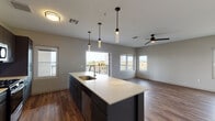 This single-floor unit showcases a contemporary design with a thoughtfully planned open-concept layout. The kitchen serves as the centerpiece, anchored by a large island with a light quartz countertop, an undermount sink, and a gooseneck faucet. Dark gray cabinetry pairs with a herringbone tile backsplash, and stainless steel appliances—including a gas range, microwave, and French door refrigerator—complete the well-equipped space. Cylindrical pendant lights hang above the island, adding a modern accent. The kitchen flows seamlessly into a generous living area finished with warm wood-look flooring, neutral gray walls, and a ceiling fan overhead. Multiple windows allow abundant natural light to fill the space, while a sliding door opens onto a private balcony enclosed by a sleek horizontal metal railing. The balcony offers elevated views of the surrounding urban landscape, including mature trees and neighboring buildings. A carpeted bedroom provides a comfortable retreat, featuring a ceiling fan and windows with additional outdoor views. The bathroom is finished with a wide quartz vanity, dark gray cabinetry, a large mirror, matte black fixtures, and a glass-enclosed walk-in shower with marble-style tile.