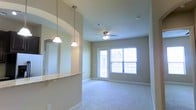 This single-floor residence offers a well-designed, open-concept layout that blends everyday functionality with comfortable living. The living area is bright and welcoming, featuring plush wall-to-wall carpeting, neutral-toned walls, and a ceiling fan that enhances air circulation throughout the space. A glass-paned door floods the room with natural light and provides direct access to an outdoor area, creating a seamless indoor-outdoor connection. The kitchen is positioned adjacent to the living space and is defined by a stylish pass-through breakfast bar topped with light granite-style countertops. Elegant pendant lights hang above the bar, adding a warm and decorative touch while maintaining an open sightline between the kitchen and living areas. The arched architectural detail framing the pass-through adds a refined design element to the overall interior. Recessed ventilation and a central ceiling fixture contribute to a practical and well-finished overhead design. Clean white baseboards trim the perimeter of each room, complementing the soft color palette throughout. Overall, the space presents a cohesive and inviting atmosphere ideal for comfortable everyday living.