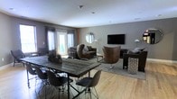 This single-floor space combines an open-concept living and dining area with a cohesive modern industrial aesthetic. The dining area anchors one side of the room with a substantial reclaimed wood table paired with contemporary black wire-frame chairs, creating a striking contrast between rustic and modern design elements. Light hardwood flooring runs throughout, adding warmth and continuity across the space.

The living area is thoughtfully arranged with a mix of seating options, including distinctive cognac leather egg chairs and a coordinating sofa, all positioned around a patterned area rug. A wall-mounted flat-screen television serves as a focal point on one end, while a round mirror adds depth and visual interest to the opposing wall. Neutral gray walls provide a calm backdrop that complements the warm tones of the furnishings.

Natural light enters generously through large French doors dressed with gray curtains, offering a view of greenery outside. Recessed ceiling lighting supplements the natural light, ensuring the space remains bright and welcoming. Potted plants are placed throughout, adding organic texture and life to the overall design.