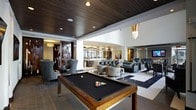 This refined single-floor amenity space blends elegant design with a social atmosphere, offering a variety of distinct zones for relaxation and entertainment. At the forefront is a striking billiards area anchored by a handsome wood-framed pool table with a dark felt surface, set beneath a rich wood-paneled ceiling with recessed lighting that creates a warm, intimate ambiance. Beyond the billiards zone, the space opens into a bright and airy lounge area featuring plush gray upholstered seating arrangements, including sofas and accent chairs, complemented by a bold area rug and contemporary art pieces adorning the walls. Pendant lighting with metallic and glass finishes adds a glamorous touch throughout. A bar and counter area is visible in the background, appointed with sleek modern fixtures and pendant lights, ideal for social gatherings. A more intimate lounge nook is tucked to one side, partially screened by a decorative partition with an art deco-inspired lattice design, offering a cozy retreat within the larger space. The interplay of dark wood, light walls, polished marble-look flooring, and curated lighting creates a cohesive, upscale atmosphere throughout this well-appointed communal space.