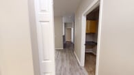 Located at 1200 Bismark Lane SE in Lacey, Washington, this single-floor unit offers a thoughtfully designed layout suited for comfortable everyday living. The open-concept living area is finished with light wood-style luxury vinyl plank flooring and is bathed in natural light through large windows fitted with horizontal blinds, with a door providing direct outdoor access. The kitchen is efficiently arranged with warm-toned wood cabinetry, dark granite-style countertops, and stainless steel appliances including a refrigerator, over-the-range microwave, and an electric range. A hallway connects the main living spaces, with a convenient built-in shelving nook visible along the corridor. The bedrooms feature plush carpeting and neutral-toned walls; one bedroom includes a mirrored sliding closet, while another offers a window with views of surrounding greenery and a baseboard heater. The unit includes two bathrooms: a full bath with a soaking tub and shower combination accented by white tile surround and a brushed nickel grab bar, and a half or secondary bath featuring a single-basin vanity with granite-style countertops, warm wood cabinetry, and light vinyl plank flooring. The overall ambiance is clean, neutral, and move-in ready.