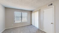 This single-floor residence offers a practical and well-organized layout designed for comfortable everyday living. The living area is carpeted and filled with natural light streaming through a large window fitted with horizontal blinds, complemented by a textured ceiling. A sliding glass door with vertical blinds provides additional light and access to the outdoors. The kitchen is efficiently arranged with white cabinetry featuring black hardware, light speckled countertops, and wood-look vinyl flooring. Black appliances, including a refrigerator and range, anchor the space, while a peninsula-style breakfast bar opens the kitchen toward the living area, creating a connected and functional flow. A second carpeted room serves as the bedroom, equipped with a ceiling fan with integrated lighting and its own door for privacy. The bathroom is clean and utilitarian, featuring a full soaking tub with a shower surround, a toilet, and overhead white cabinetry with black hardware for additional storage. Throughout the unit, neutral warm-gray tones on the walls, textured ceilings, and consistent trim work create a cohesive and understated aesthetic. The layout is straightforward and efficient, making good use of the available space.