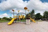 This outdoor recreational space centers around a well-equipped, nature-themed playground structure designed to engage children of varying ages and abilities. The play structure features a distinctive design highlighted by tall, mushroom-capped poles in bold yellow and green tones that give the area a whimsical, organic aesthetic. Two yellow slides extend from the main platform, offering exciting descent options, while a leaf-patterned climbing wall provides an alternative route to the top. Suspended yellow play elements beneath an arched frame add an additional interactive challenge for climbers and adventurers alike. The entire structure is set on a generous gravel surface that provides safe, impact-absorbing ground cover throughout the play area. A wooden bench nearby offers seating for supervising adults. The surrounding landscape features open green lawns and mature trees that frame the space and provide natural shade. In the background, a fenced pool area is visible, suggesting this playground is part of a larger recreational complex. A small storage room on-site includes shelving, hanging storage, and cabinetry, supporting the operational needs of the facility. Overall, the space offers a lively and well-maintained environment for outdoor play and community recreation.